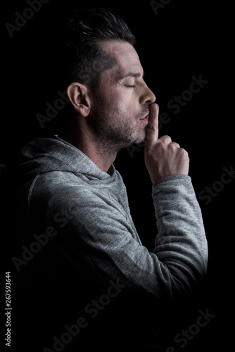 Side studio portrait of a man with closed eyes, with finger on lips making silence gesture. Isolated on black background. Vertical.
