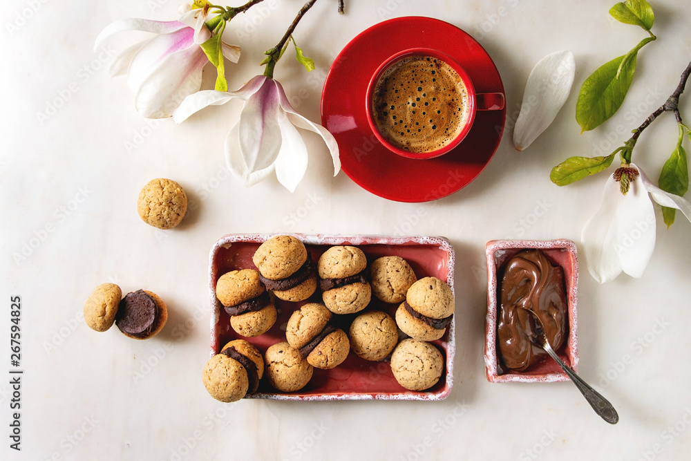 Baci di dama homemade italian hazelnut biscuits cookies with chocolate cream served in ceramic plate with red cup of espresso coffee and magnolia flowers over white marble background. Flat lay, space Baci di dama homemade italian hazelnut biscuits cookies with chocolate cream served in ceramic plate with red cup of espresso coffee and magnolia flowers over white marble background. Flat lay, space