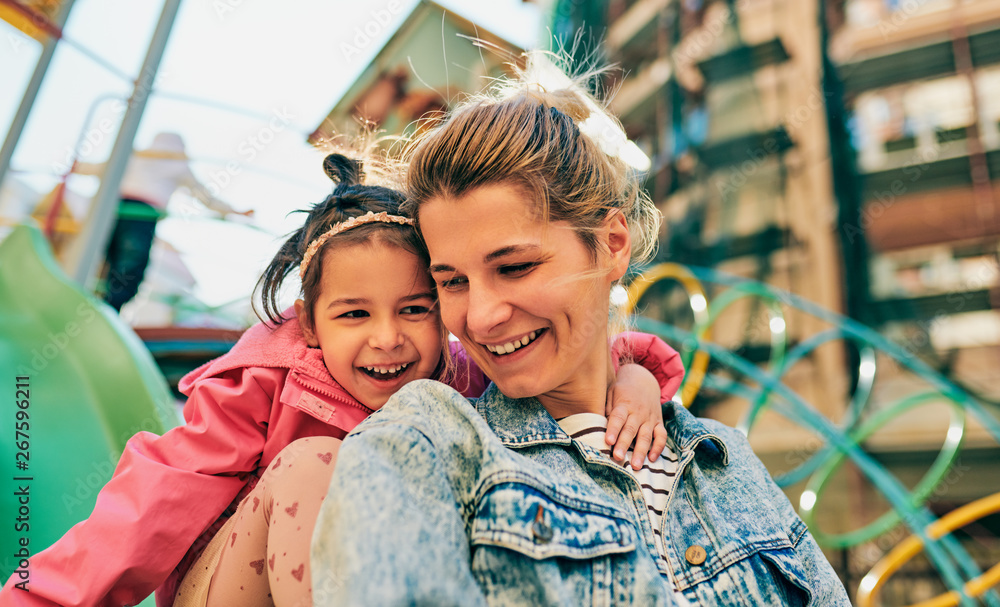 Obraz premium Outdoor portrait of happy cute little girl smiling and looking to her beautiful mother spending time together at playground. Young woman and daughter feeling happy, loving each other outside.