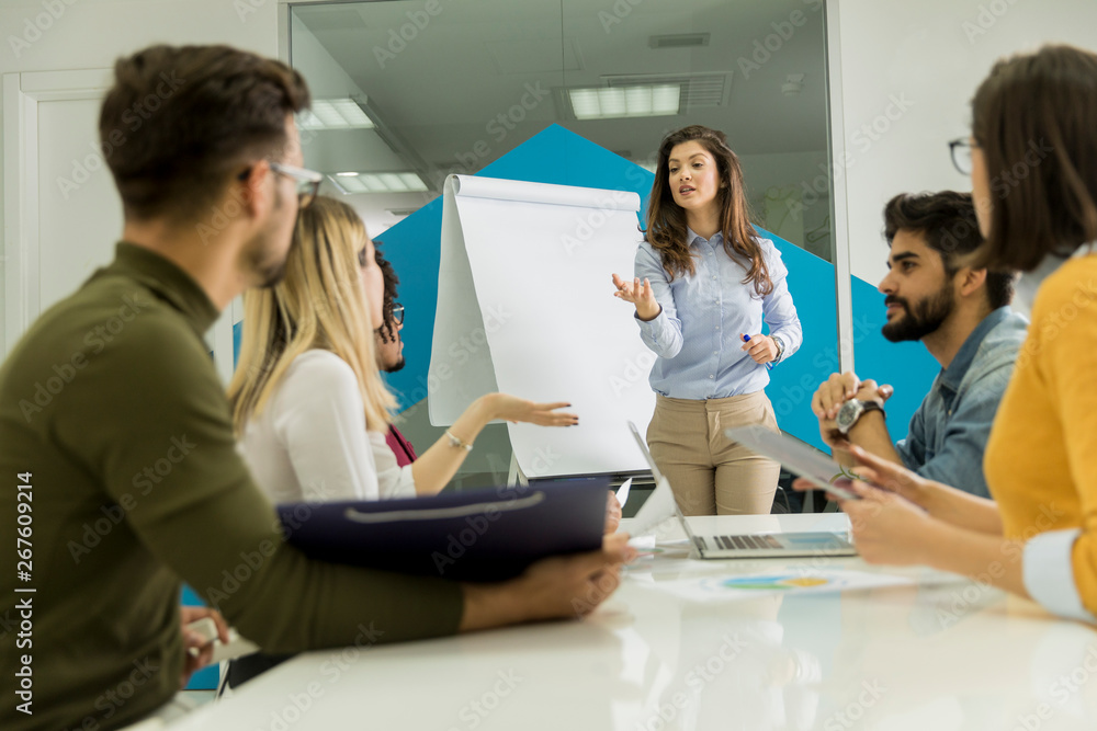Confident young team leader giving a presentation to a group of young colleagues as they sit grouped by the flip chart in the office Confident young team leader giving a presentation to a group of young colleagues as they sit grouped by the flip chart in the office