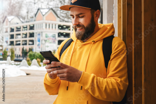 Front view. Bearded male tourist in yellow hoodie and cap with black backpack stands outdoors and using smartphone. Lifestyle.