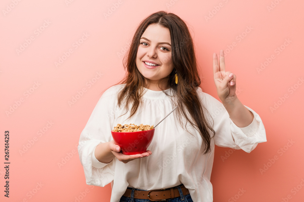 Young plus size curvy woman holding a cereals bowl showing victory sign and smiling broadly. Young plus size curvy woman holding a cereals bowl showing victory sign and smiling broadly.