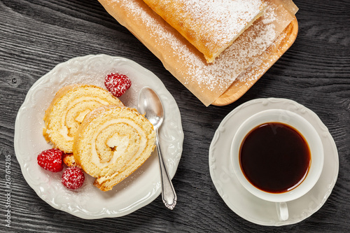 Ciasto - rolada biszkoptowa z kremem mascarpone na talerzyku, obok kawa w filiżance, wszystko postawione na czarnym drewnianym blacie photo