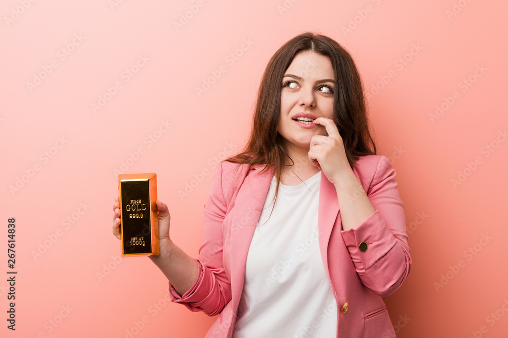 Young plus size curvy business woman holding a gold ingot relaxed thinking about something looking at a copy space. Young plus size curvy business woman holding a gold ingot relaxed thinking about something looking at a copy space.