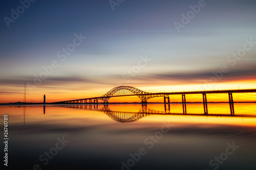 Long silhouetted bridge with an arch crossing a crystal clear calm body of water at sunset. Perfect reflections in the water - Long Island New York.   photo