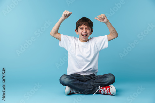 Image of happy brunette boy 10-12y with freckles wearing white casual t-shirt smiling at camera while sitting on floor