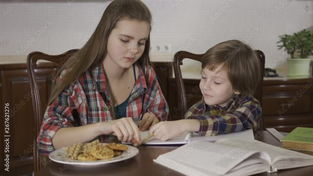 Older sister and younger brother doing homework together sitting at the table at home. The teenage girl helping little boy with math, counting and writing the correct answer. Loving friendly family Older sister and younger brother doing homework together sitting at the table at home. The teenage girl helping little boy with math, counting and writing the correct answer. Loving friendly family