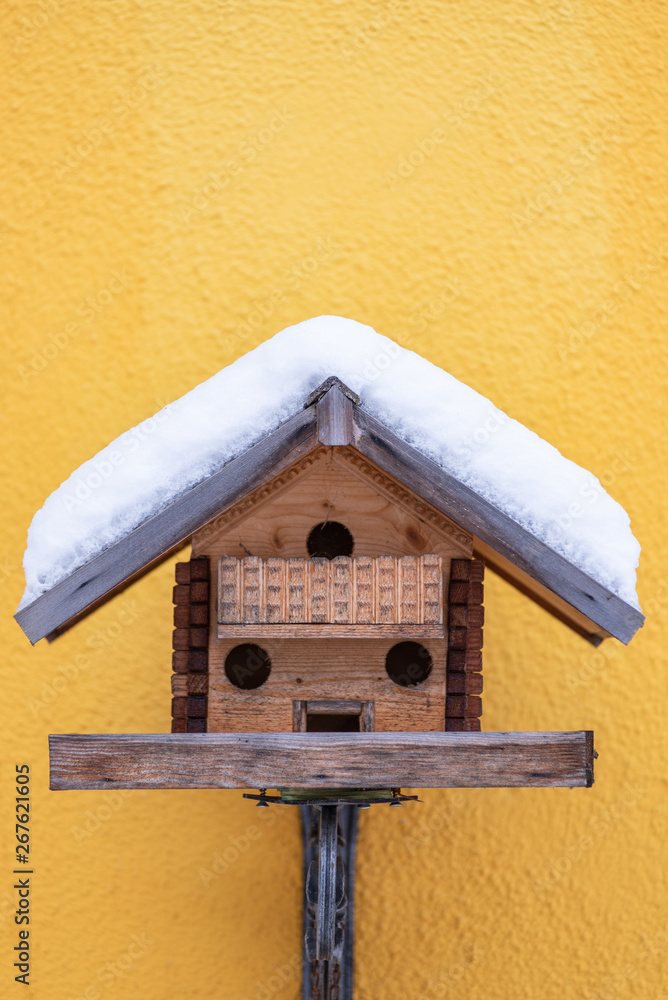 Homemade wooden bird's feeder in winter, under the snow on the yellow wall background.