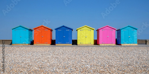 a line of six multicoloured beach huts on Seaford beach, in the foreground is a pebble beach in the back ground is a clear blue sky