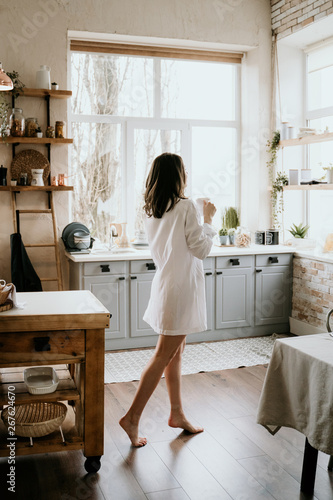 Successful and confident woman on a morning coffee break, resting and enjoying the drink. Young girl in white panties and long shirt with naked shoulder and chest stand on the kitchen of the house