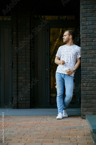 A man stands on the porch of a brick building and drinks coffee from a paper Cup in the morning