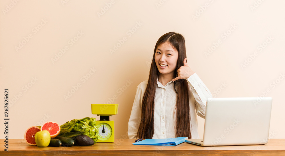 Young nutritionist chinese woman working with her laptop showing a mobile phone call gesture with fingers. Young nutritionist chinese woman working with her laptop showing a mobile phone call gesture with fingers.