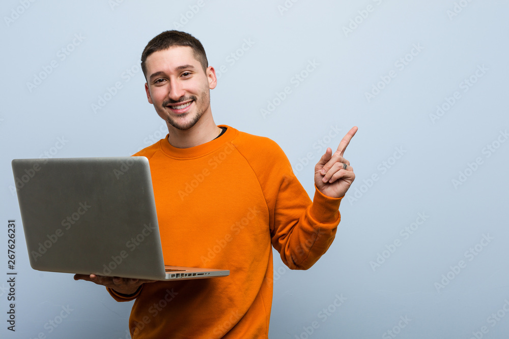 Young caucasian man holding a laptop smiling cheerfully pointing with forefinger away. Young caucasian man holding a laptop smiling cheerfully pointing with forefinger away.