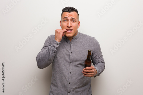 Young latin man holding a beer biting nails, nervous and very anxious