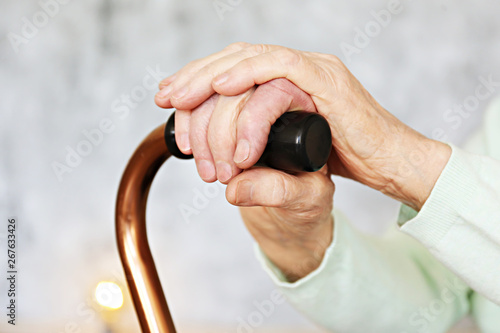 Elderly woman sitting in nursing home room holding walking quad cane with wrinked hand. Old age senior lady wearing beige cardigan, metal aid stick handle bar close up. Interior background, copy space photo