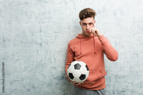 Young sporty man holding a soccer ball pointing his temple with finger, thinking, focused on a task.
