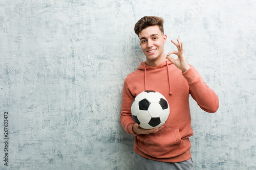 Young sporty man holding a soccer ball cheerful and confident showing ok gesture.