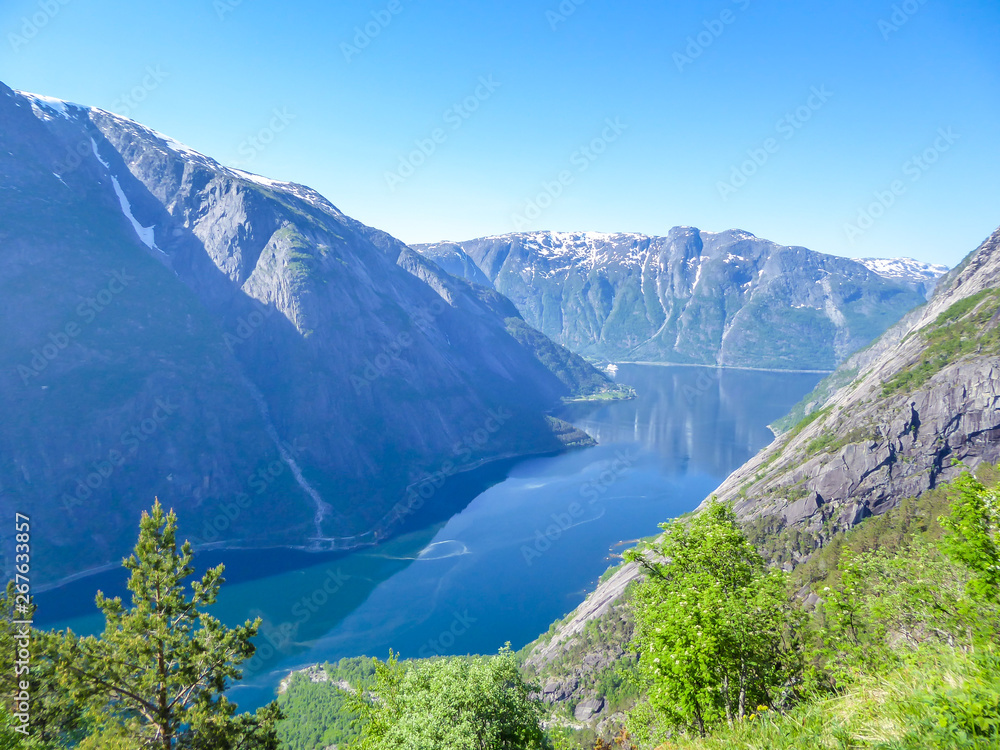 An majestic view on Eidfjord from Kjeasen, Norway. Slopes of the mountains are overgrown with lush green grass. Water has dark blue color. Taller parts of the mountains are barren. Sunny and clear day An majestic view on Eidfjord from Kjeasen, Norway. Slopes of the mountains are overgrown with lush green grass. Water has dark blue color. Taller parts of the mountains are barren. Sunny and clear day