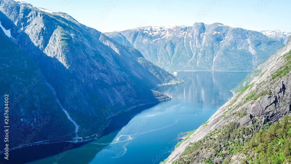 An majestic view on Eidfjord from Kjeasen, Norway. Slopes of the mountains are overgrown with lush green grass. Water has dark blue color. Taller parts of the mountains are barren. Sunny and clear day