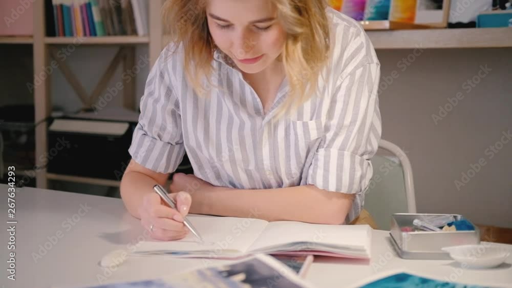 Woman artist looking at smartphone screen sketching. Neat workplace. Young blonde female smiling creating artwork. Woman artist looking at smartphone screen sketching. Neat workplace. Young blonde female smiling creating artwork.