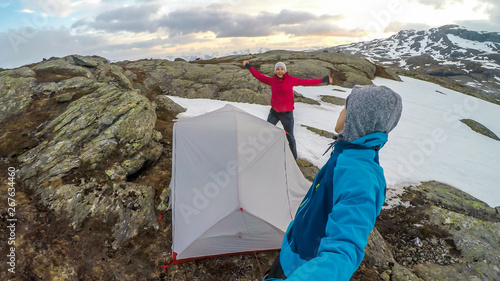 A young couple putting the tent up in the wilderness. The ground is covered with snow. Girl jumps behind the tent. Cold and windy weather. Camping in the unfavorable weather conditions. photo