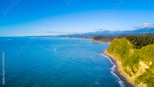 Remote rocky coastline with native forest and snowy mountain peaks on the background. West Coast, South Island, New Zealand © Dmitri