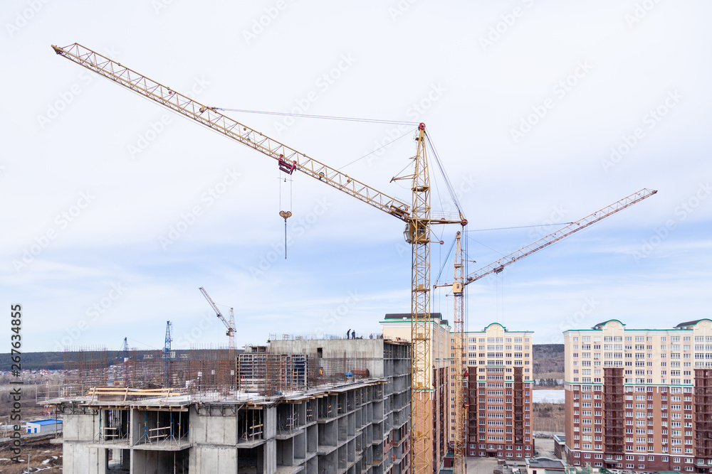 High-rise mighty hoisting crane and large building construction site with cranes with long yellow arrow against blue sky new multi-storey concrete brick, industrial background