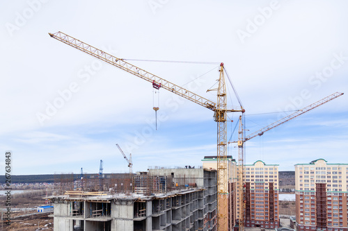 High-rise mighty hoisting crane and large building construction site with cranes with long yellow arrow against blue sky new multi-storey concrete brick, industrial background