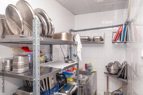 View of restaurant's professional washer, sink, brushes, metal shelving and shelves with kitchen utensils, cups, cutting board, bowls, lid, buckets, gastronomy container against white tiled wall