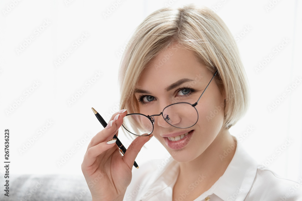 Young beautiful blonde girl with short hair in a white shirt and glasses close-up holds a pen in her hands. Holds glasses with his hands and looks out from under eyeglasses. Young beautiful blonde girl with short hair in a white shirt and glasses close-up holds a pen in her hands. Holds glasses with his hands and looks out from under eyeglasses.