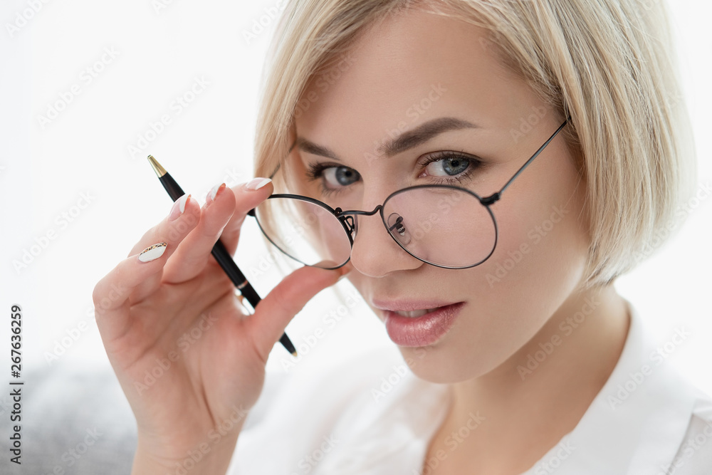 Young beautiful blonde girl with short hair in a white shirt and glasses close-up holds a pen in her hands. Holds glasses with his hands and looks out from under eyeglasses. Young beautiful blonde girl with short hair in a white shirt and glasses close-up holds a pen in her hands. Holds glasses with his hands and looks out from under eyeglasses.