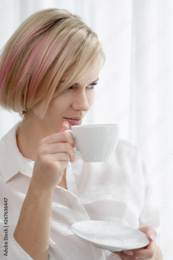 Young beautiful blonde girl with short hair in a white shirt and glasses is sitting on the sofa in bright in the office against the window. Drinking coffee or tea in a working break. Young beautiful blonde girl with short hair in a white shirt and glasses is sitting on the sofa in bright in the office against the window. Drinking coffee or tea in a working break.