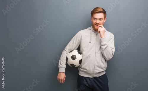 Young redhead fitness man relaxed thinking about something looking at a copy space. He is holding a soccer ball.