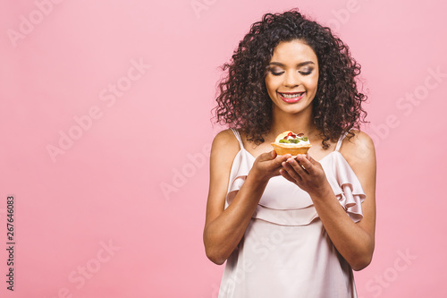 Black american african happy woman with curly afro hair style making a mess eating a huge fancy dessert over pink background. Eating cupcake.