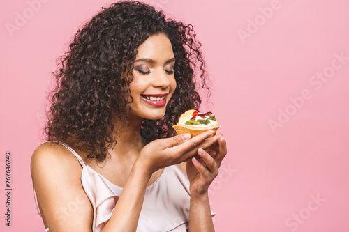 Black american african happy woman with curly afro hair style making a mess eating a huge fancy dessert over pink background. Eating cupcake.