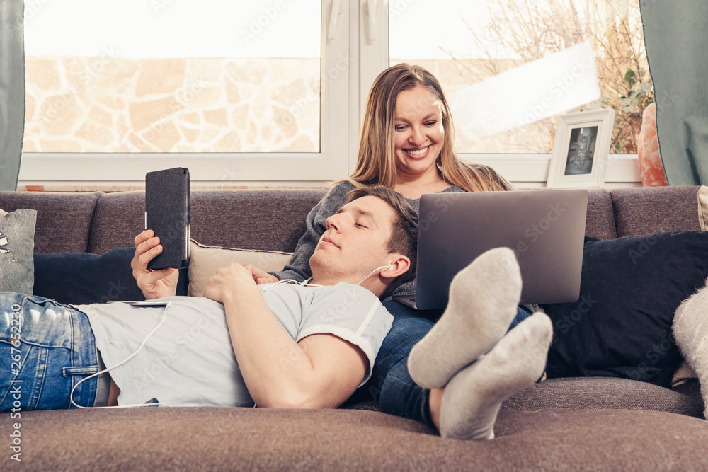 Young couple relaxing on sofa and using notebook, smartphone and ebook reader Young couple relaxing on sofa and using notebook, smartphone and ebook reader