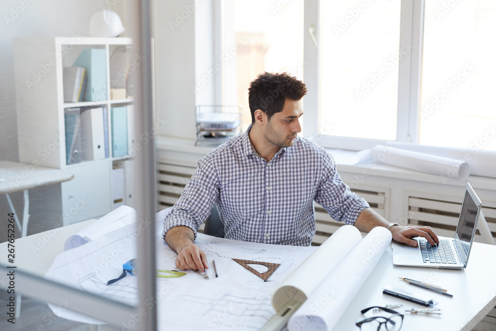 Portrait of young engineer working with plans and blueprints sitting at desk in office