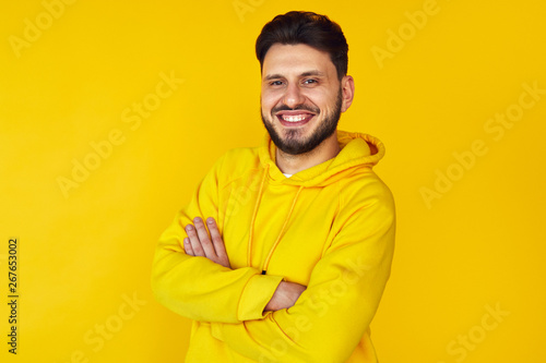 Young hipster male in yellow hoodie smiling and posing with arms crossed over yellow background