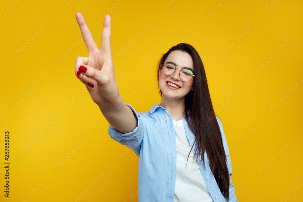 Beautiful young female with eyeglasses dressed in blue shirt and showing peace gesture isolated over orange background Beautiful young female with eyeglasses dressed in blue shirt and showing peace gesture isolated over orange background