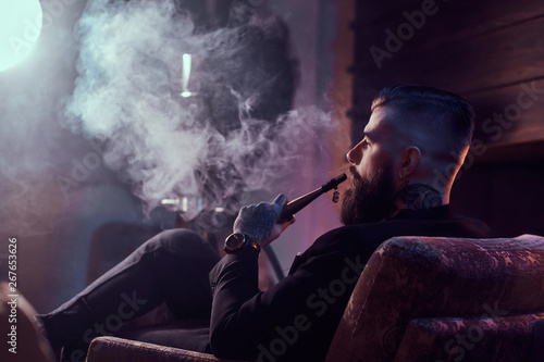 Attractive bearded man is making nice misty vapour while relaxing near fireplace and smoking hookah.