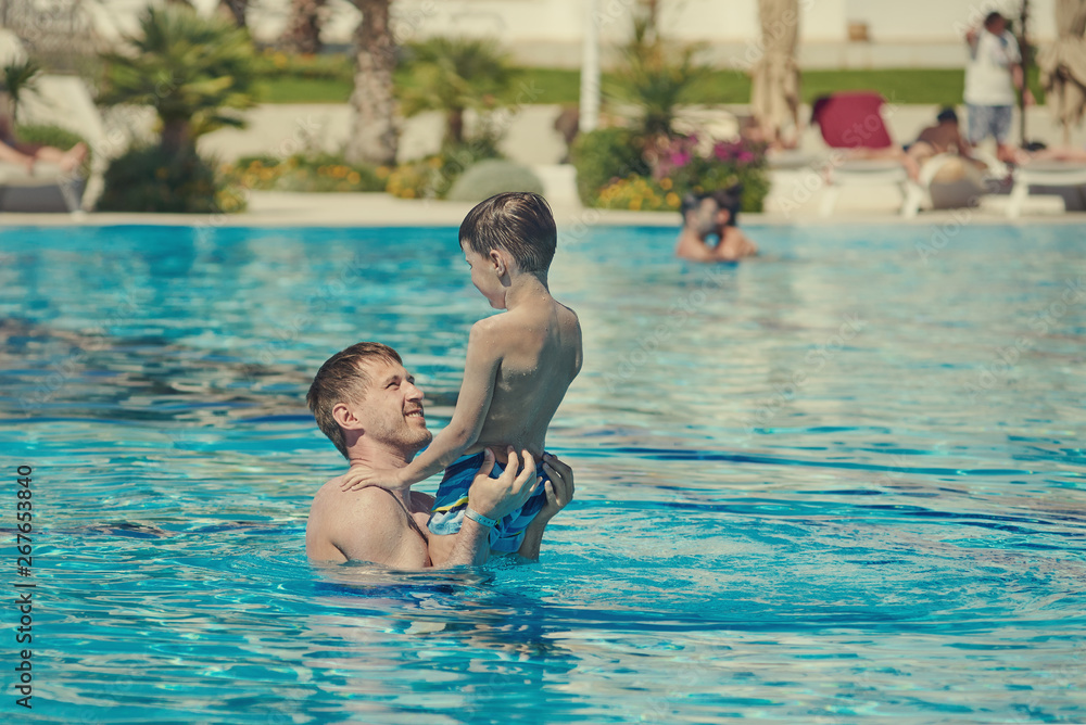 Dad is holding his son in his arms, while having fun in the luxurious pool.