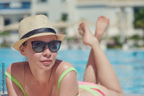 Young beautiful European woman in a sunhat and sunglasses is laying on the sunbed near the hotel’s swimming pool, she is enjoying her holidays.