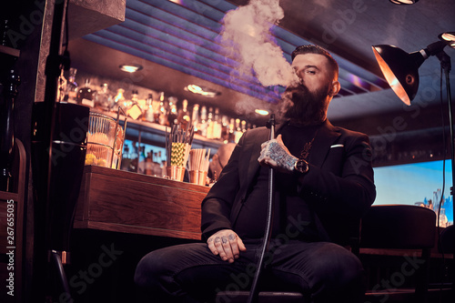 Portrait of tattooed bearded man which is smoking hookah, making nice vapour while sitting near bar counter.