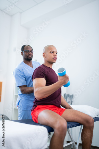 Dark-skinned therapist in glasses watching his patient doing exercises