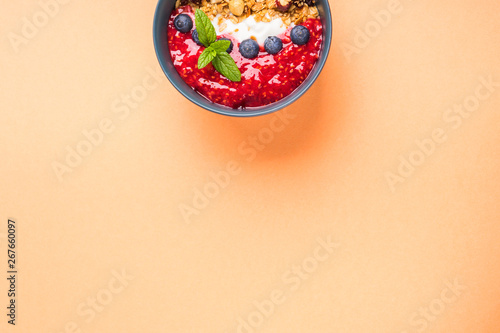 Yogurt smoothie bowl with blended raspberries, blueberries and oat matcha granola on cantaloupe orange pastel background. Delicious healthy breakfast concept