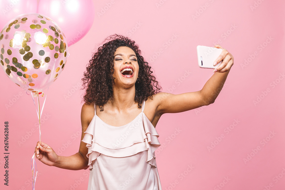Self portrait of afro american positive girl in dress having balloons in hand shooting selfie on front camera isolated on pink background. Self portrait of afro american positive girl in dress having balloons in hand shooting selfie on front camera isolated on pink background.