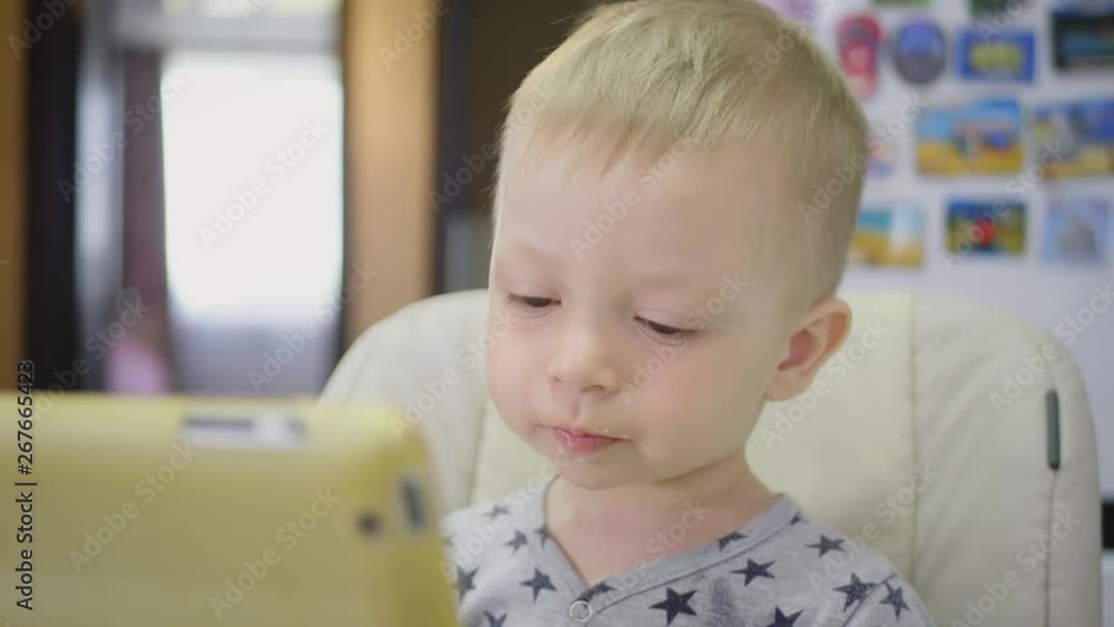 Cute little boy, eating porridge for breakfast at home, while watching cartoon on tablet. Children and technology or healthy food for kid concept. Cute little boy, eating porridge for breakfast at home, while watching cartoon on tablet. Children and technology or healthy food for kid concept.