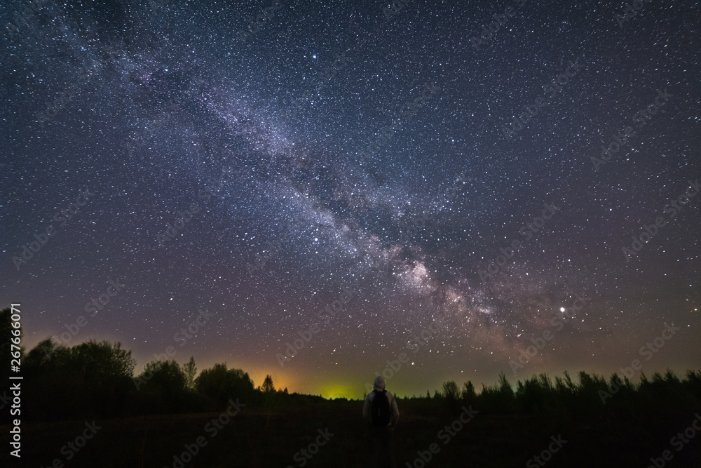 A man with a backpack standing at night, with his back to the camera, against the background of the Milky Way