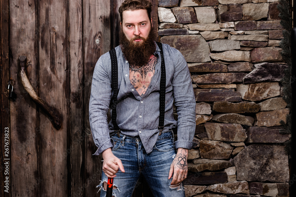 Brutal strong man with a beard and tattoos on his hands dressed in stylish casual clothes stands on the background of stone wall and wooden door