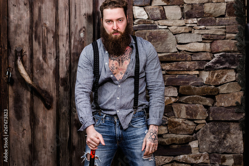 Brutal strong man with a beard and tattoos on his hands dressed in stylish casual clothes stands on the background of stone wall and wooden door